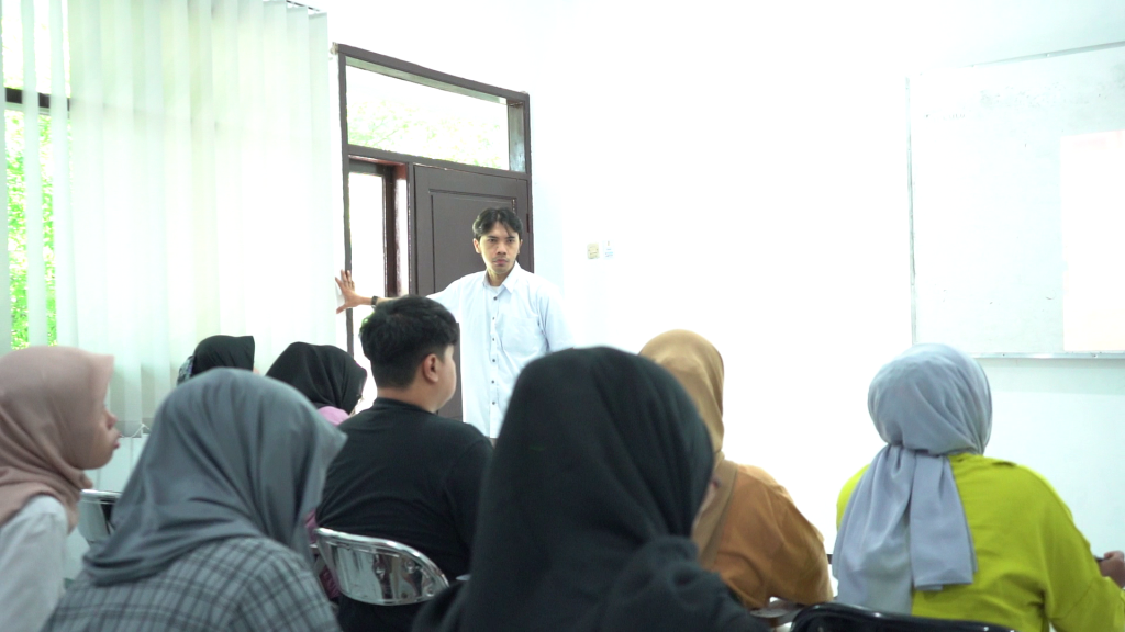 Suasana presentasi atau belajar mengajar di mana seorang pria berbicara di depan sekelompok audiens di dalam ruangan kelas yang terang.