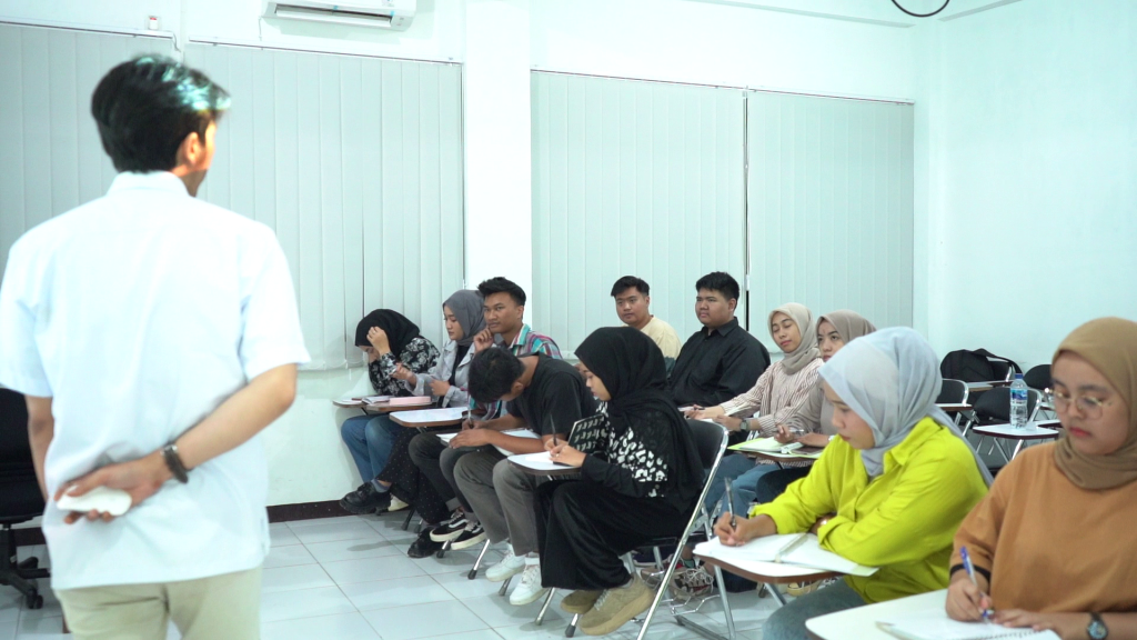 Suasana perkuliahan kelas karyawan di mana seorang dosen sedang mengajar di depan kelas, sementara para mahasiswa dewasa dengan latar belakang beragam tampak fokus memperhatikan dan mencatat materi.