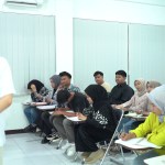 Suasana perkuliahan kelas karyawan di mana seorang dosen sedang mengajar di depan kelas, sementara para mahasiswa dewasa dengan latar belakang beragam tampak fokus memperhatikan dan mencatat materi.