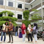 Foto sekelompok mahasiswa universitas yang ceria sedang berdiskusi santai di halaman terbuka dekat gedung kampus modern, mencerminkan dinamika belajar dan kehidupan kampus bagi mahasiswa jurusan manajemen.