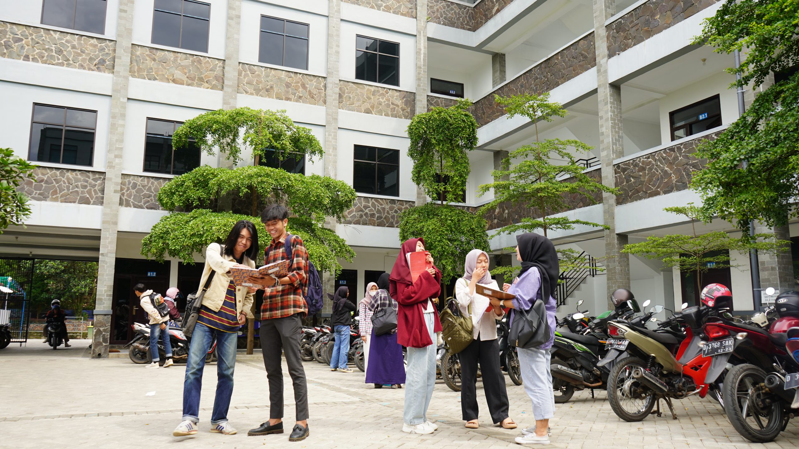 Foto sekelompok mahasiswa universitas yang ceria sedang berdiskusi santai di halaman terbuka dekat gedung kampus modern, mencerminkan dinamika belajar dan kehidupan kampus bagi mahasiswa jurusan manajemen.